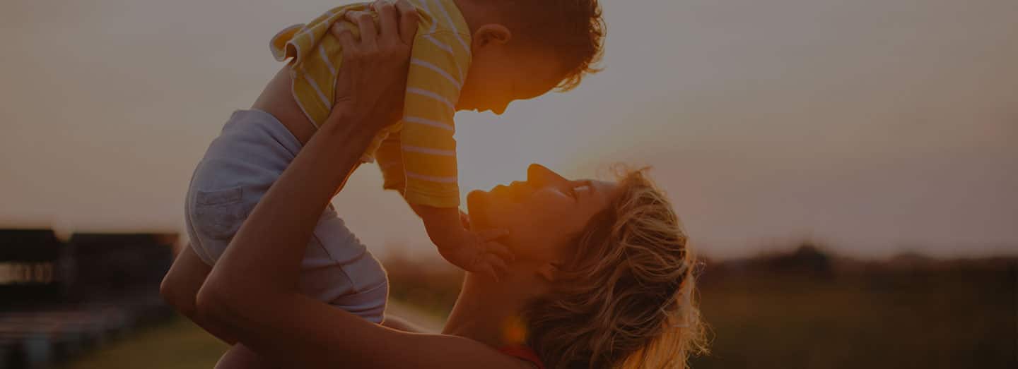A woman is holding up a small child in front of the sunset. The sunset is peaking through them as they are smiling at each other.