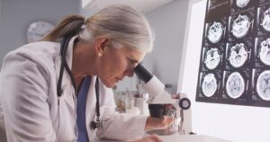 A female researcher with a silver ponytail looks through a microscope.