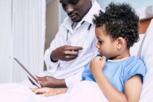 Doctor sits with little boy in the hospital bed, showing him information on a tablet.