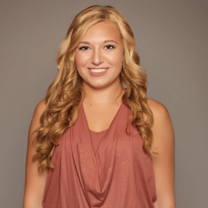 Calista Hall stands in front of a brown background, smiling in a stylish pink top and dark jeans.in a