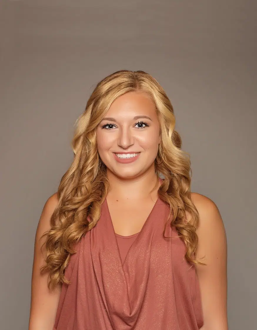 Calista Hall stands in front of a brown background, smiling in a stylish pink top and dark jeans.in a