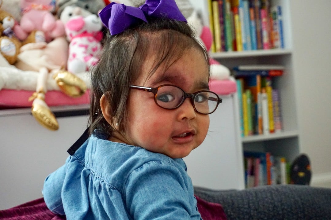 Two-year-old Vera looks back at the camera, glasses askew. She is wearing a blue hoodie and as a bow on her head.