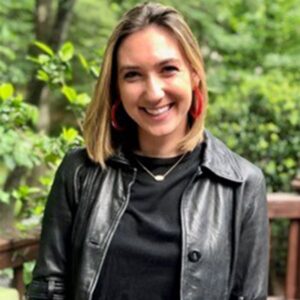 Maria stands in the sunshine wearing a nice black jacket over a black shirt.
