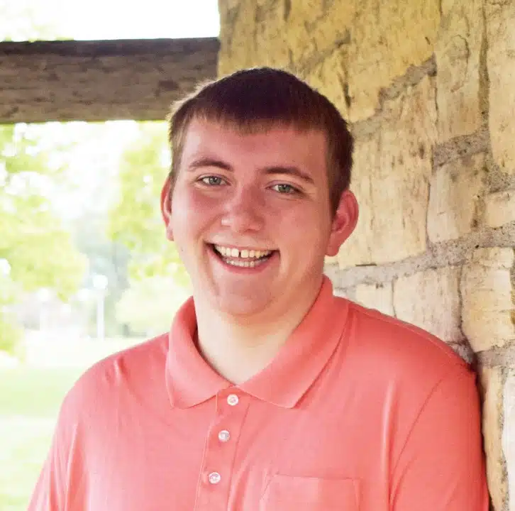 Trevor Phillips wears a salmon colored shirt and leans against a light brown brick wall.