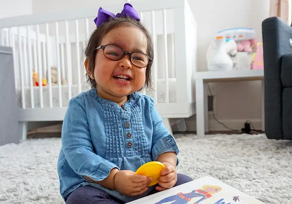 Two-year-old Vera is sitting in her bedroom. She is all smiles and is wearing a blue shirt and purple hair bow.