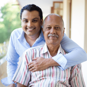 a younger man hugs his elderly father from behind as they smile at the camera