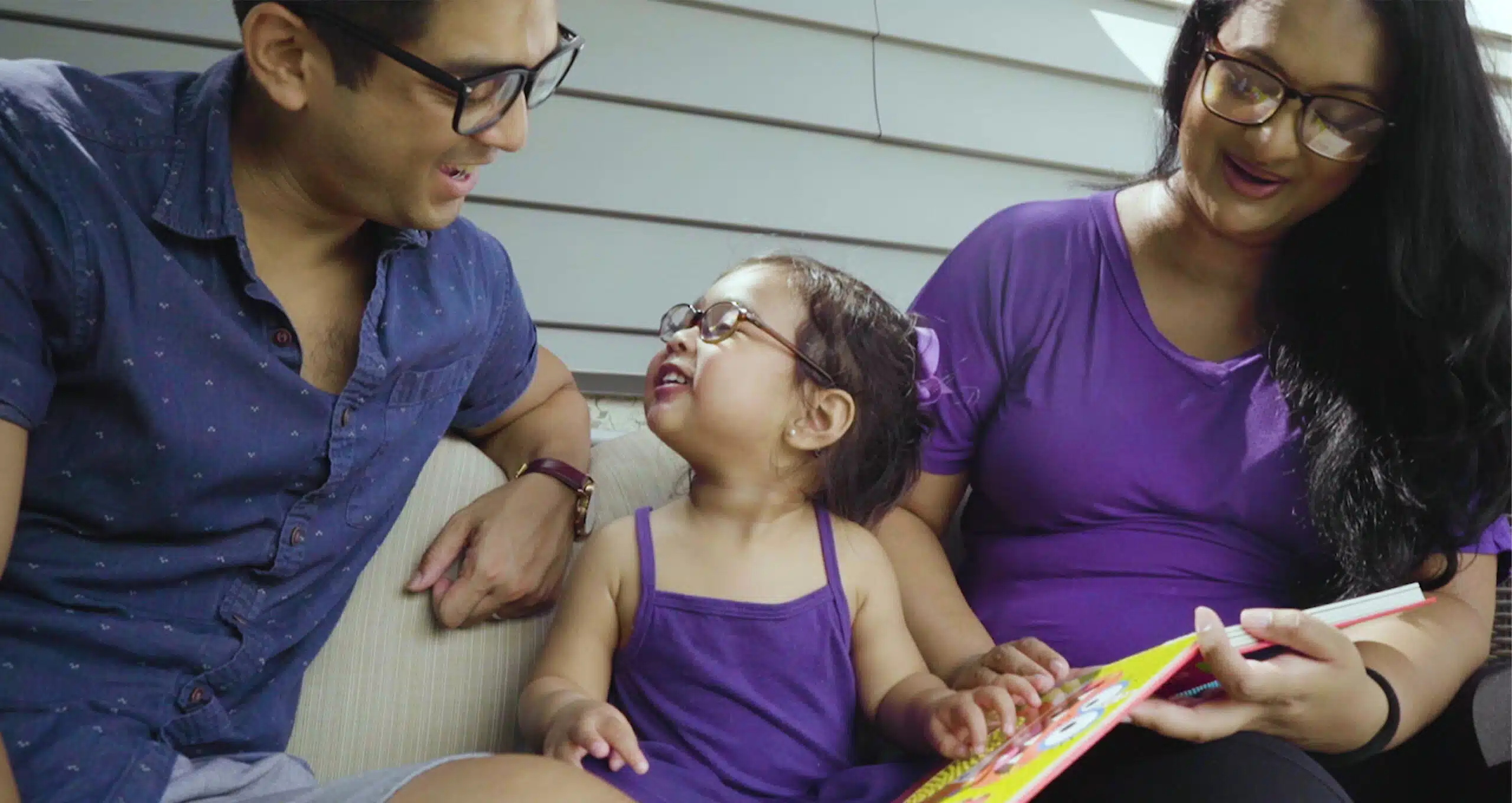 Parents and their child with epilepsy seated outside laughing and reading a book