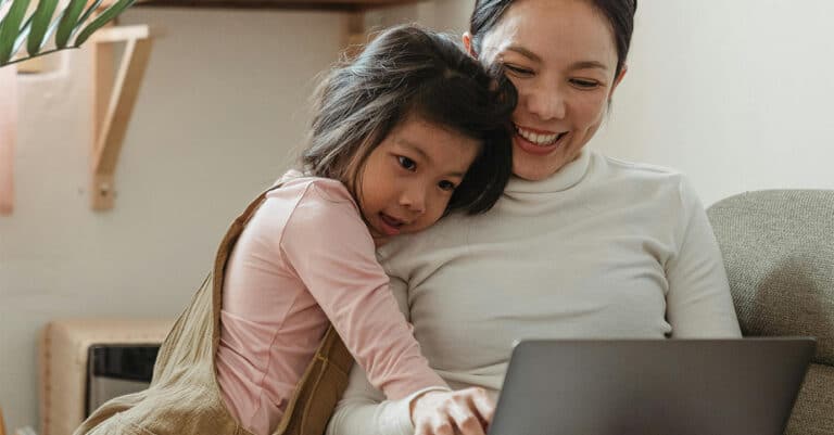 An adult and child sitting together looking at a laptop, with a light background and sofa.