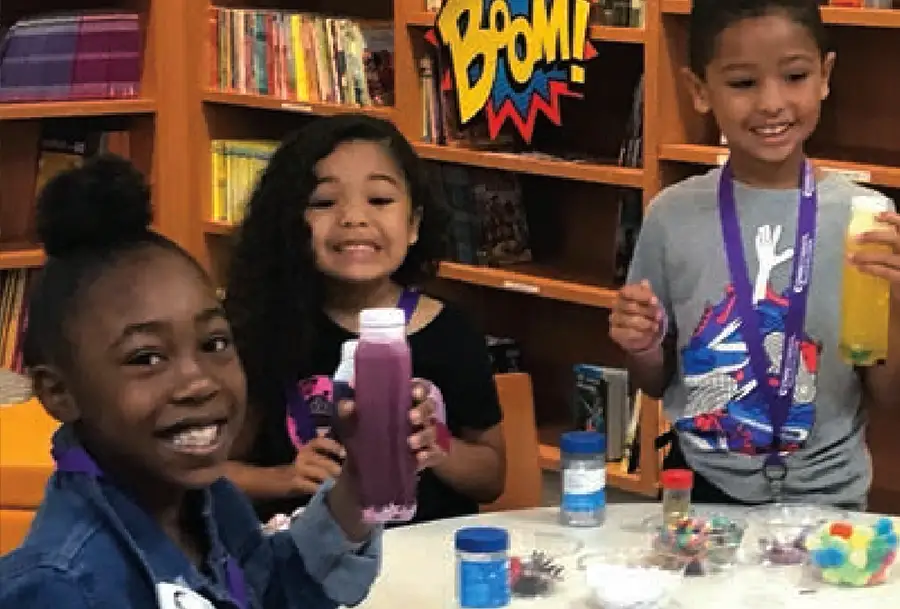 Photo of 3 children holding drinks in front of table in classroom-like setting