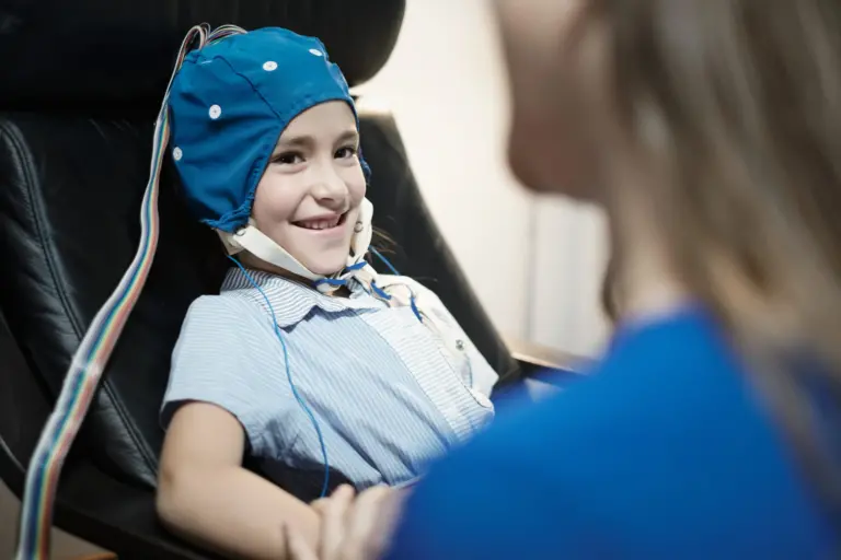A child is hooked up to an EEG test is smiling, holding hands with the woman across from him.