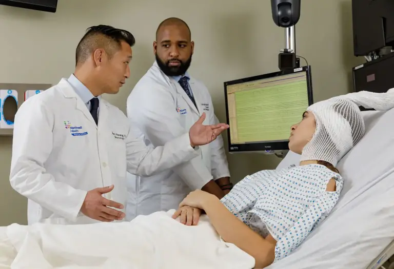 Two doctors discuss epilepsy treatment options in front of a patient in a hospital bed hooked up to EEG wires
