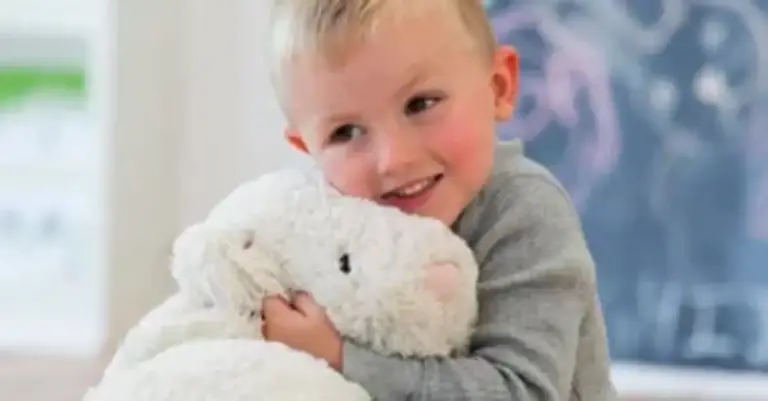 A small child is hugging a teddy bear, smiling, in a brightly lit room.
