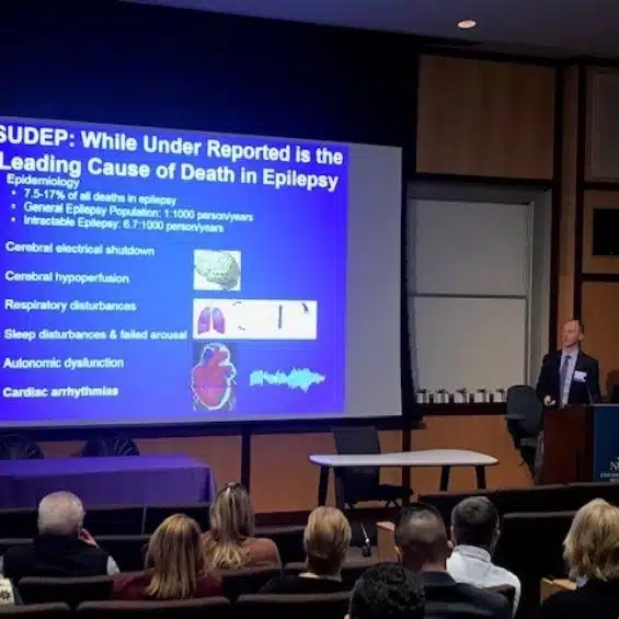 A man standing at a podium giving a presentation titled SUDEP: While Under Reported is the Leading Cause of Death in Epilepsy.