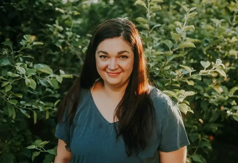 Photo of woman wearing green shirt smiling at camera in front of a greenery background.
