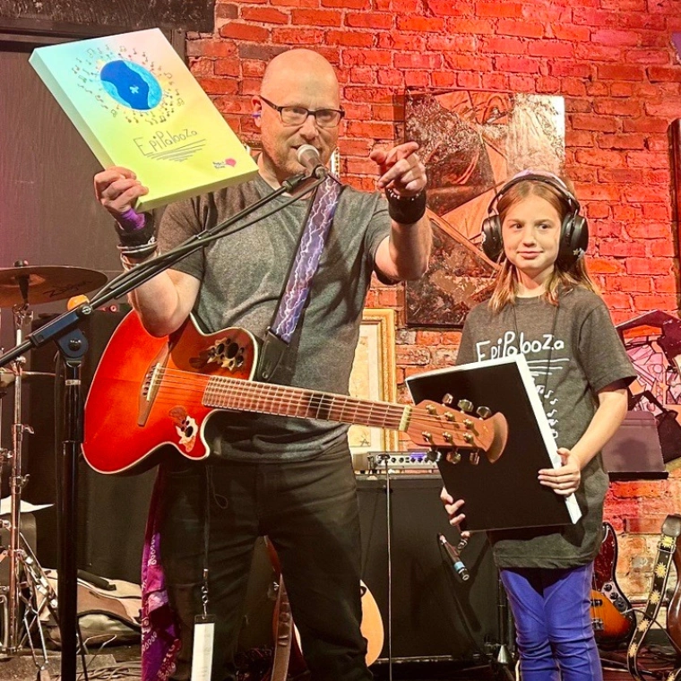 Matt Perrone onstage with a child wearing a guitar and holding a book in one hand while speaking into a microphone and pointing to the crowd.