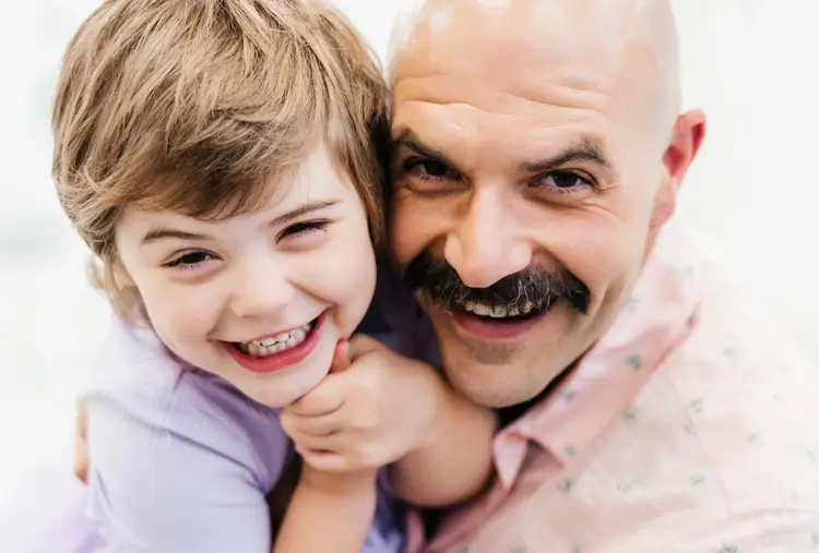 Photo of a father and son hugging and smiling at the camera up close in a brightly lit setting.