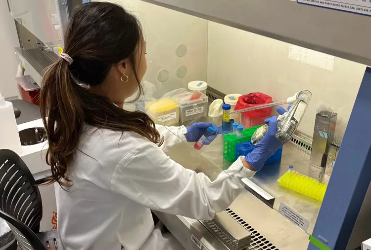 A person in a lab coat working at a laboratory bench with pipette and colored containers.