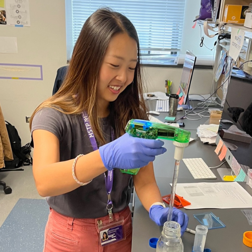 A young researcher is working in a lab. She is wearing blue gloves and is manipulating a jar of fluid.