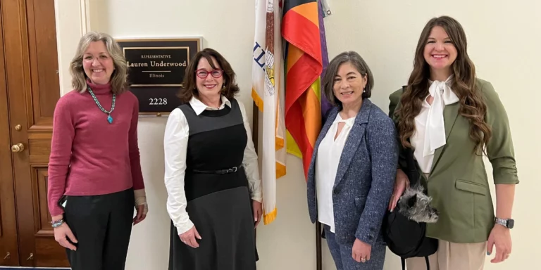 National Plan Hall Photo of four women stand in front of a wall with a plaque and flags.