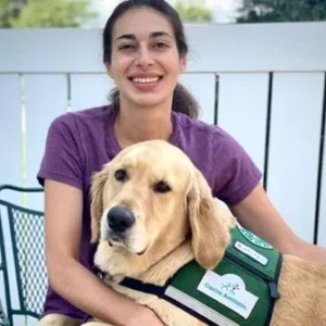 Channing is a young woman and is posing with her service dog, Bishop. Channing is wearing purple and smiling.