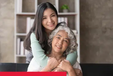 An elderly mother sits in front of her daughter, who is hugging her from behind. They are looking into the camera and smiling.