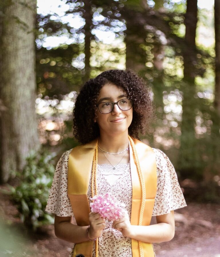Photo of a young woman in graduation sash holding flowers and smiling.