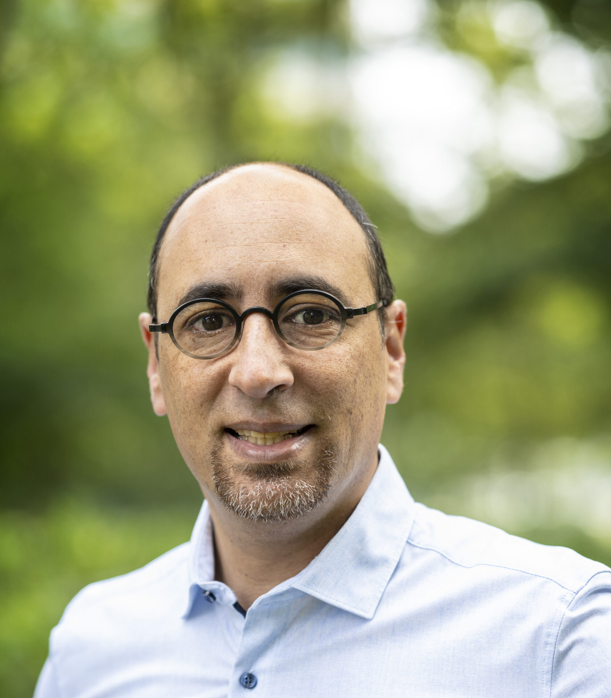 Man wearing glasses and blue dress shirt smiles at camera with greenery in the background.