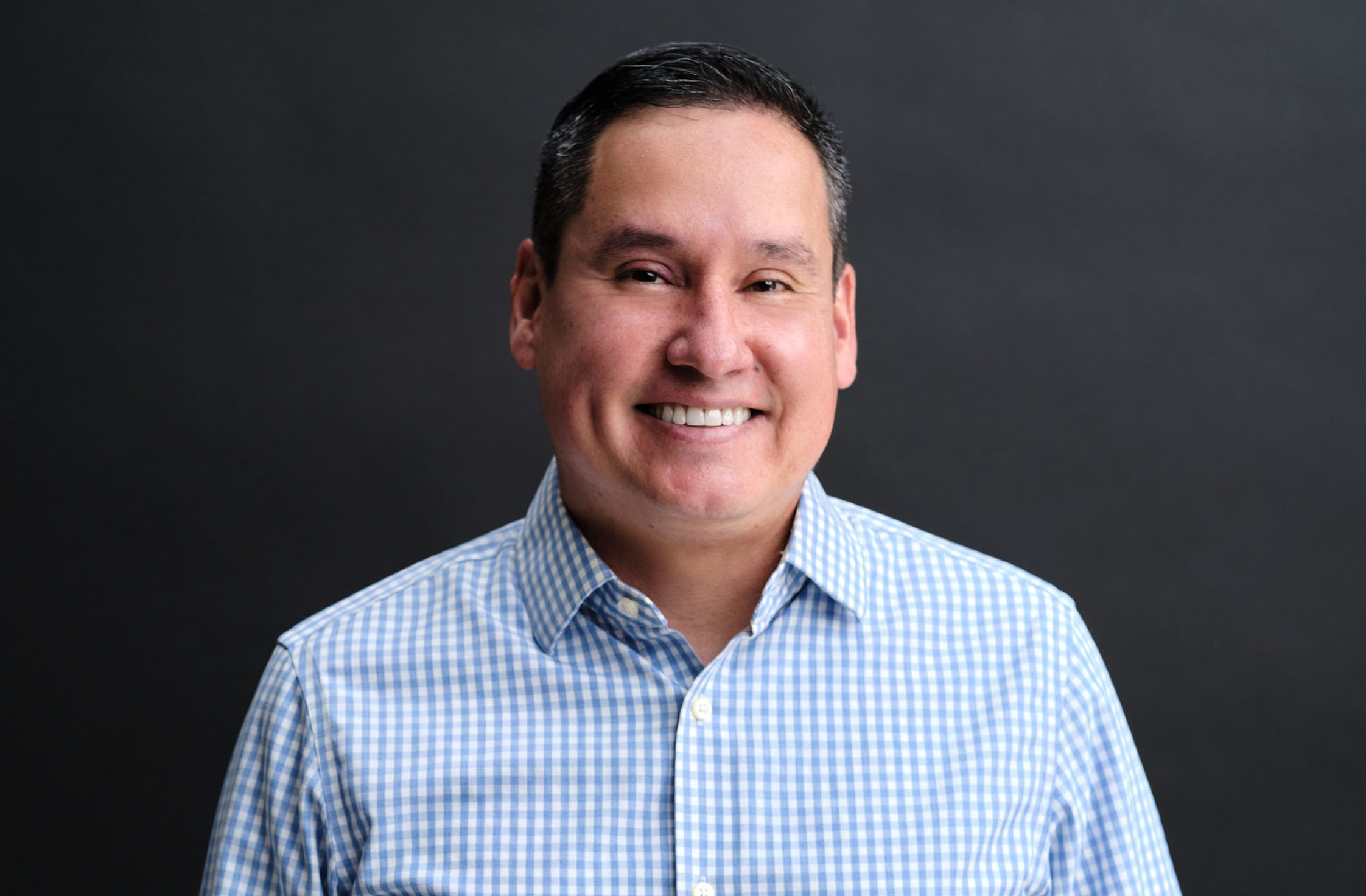 Luis Williams smiles at camera for headshot wearing a blue dress shirt standing in front of a black background.
