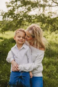 Mom Amber has her arms lovingly around grade-schooler Max. They're wearing white shirts and standing in a sunny field.