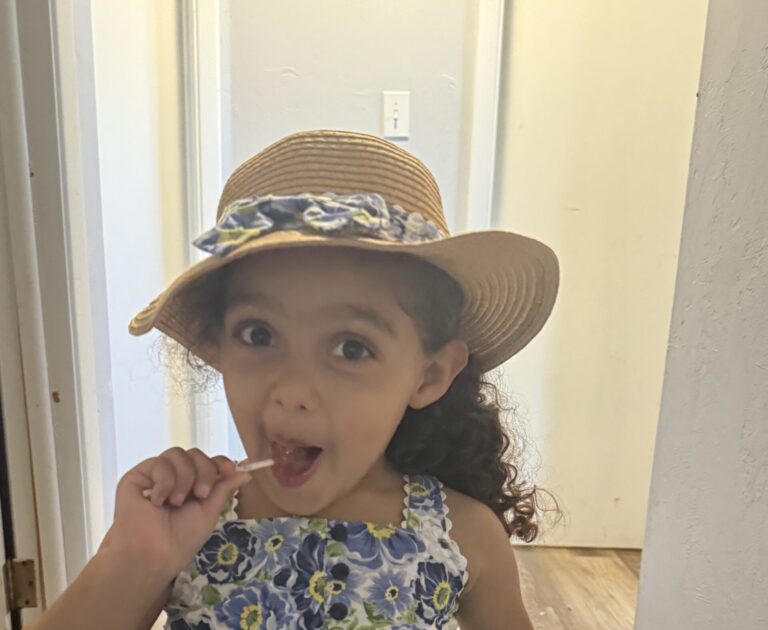 A little girl wearing a floral dress and sun hat smiles at camera holding candy to her mouth.