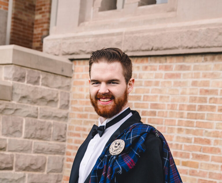 A man wearing a kilt smiles at the camera in front of a brick building.
