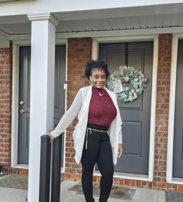 Photo of woman standing in front of a house wearing red shirt and a white cardigan smiles at the camera.