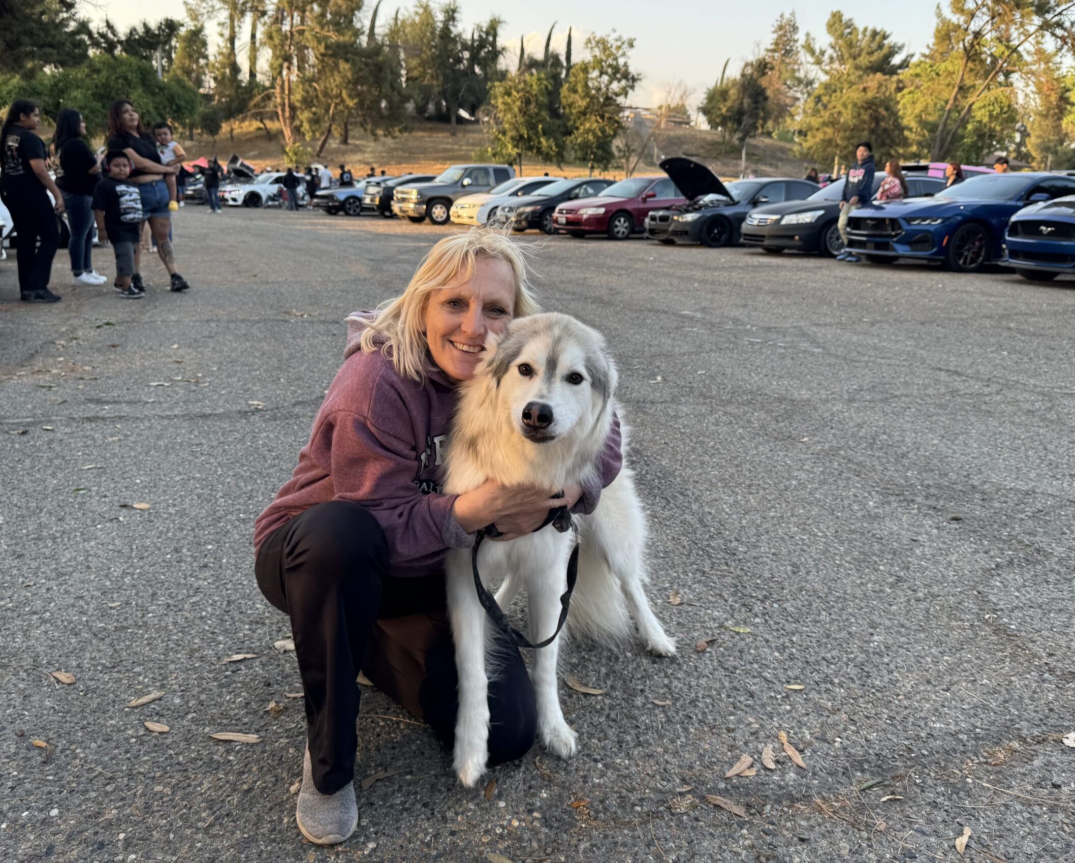 A woman smiles next to her dog in a parking lot outdoors.