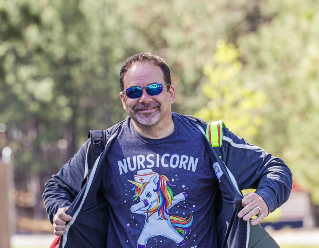 Man smiling, wearing sunglasses and showing off his epilepsy related unicorn t shirt outdoors in front of greenery.