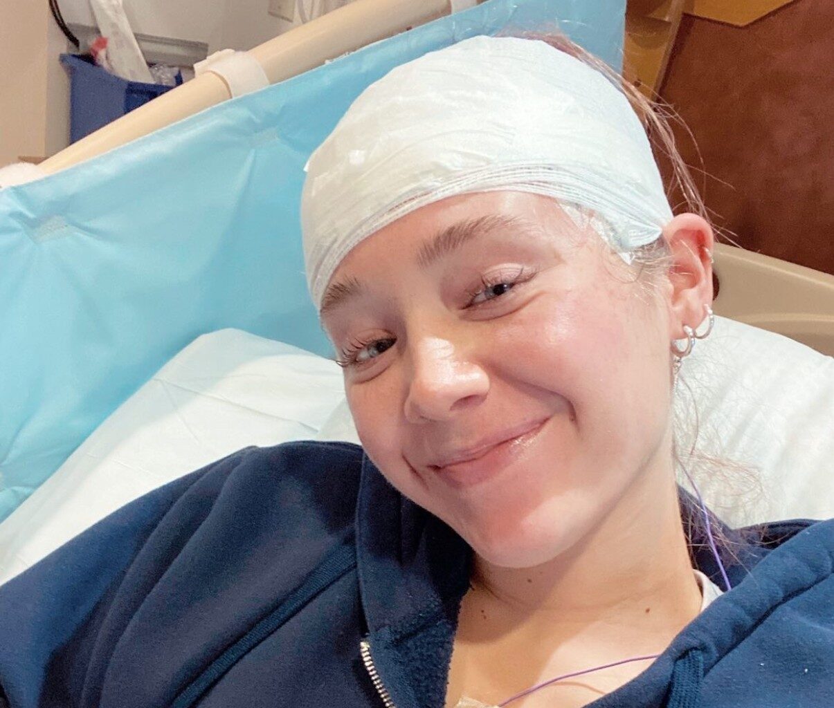 A young woman with epilepsy smiles for a selfie in a hospital bed wearing EEG headband.