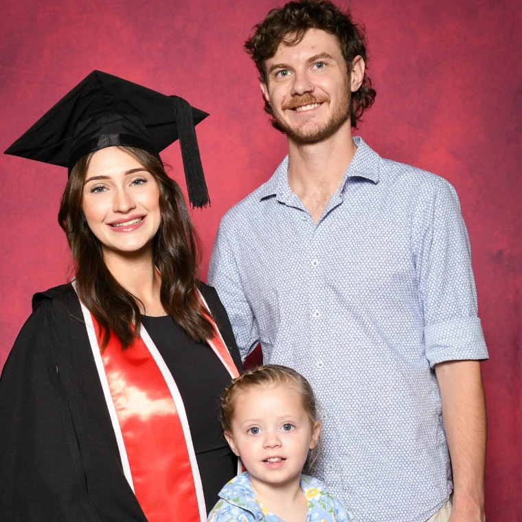 Family smiling in front of red background. The mother is wearing graduation cap and gown next to her husband and daughter pictured in the middle.