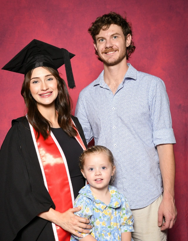 A family is pictured standing closely in front of a red background while the mother wears graduation cap and gown.