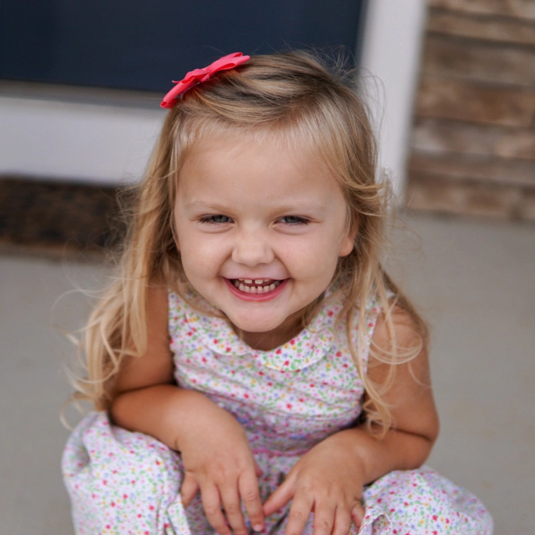 Young girl smiles at camera crouching down in a dress with her hands in lap.