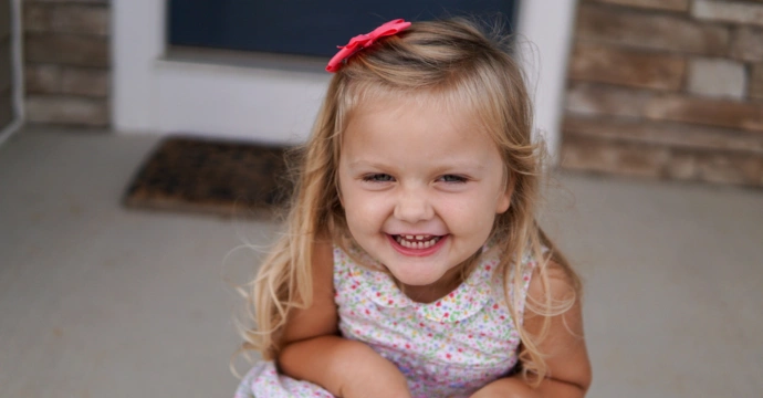 A young girl smiles at the camera wearing a bow in her hair and crouching down in her dress pictured on a porch.