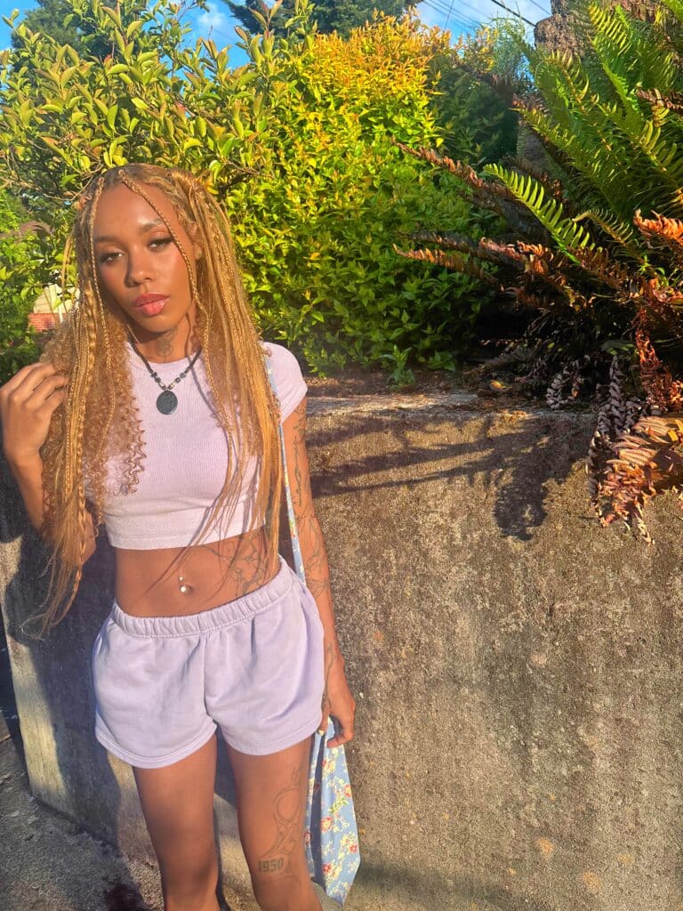 A young woman poses for a photo wearing a white top and pink shorts in front of a luscious green background.