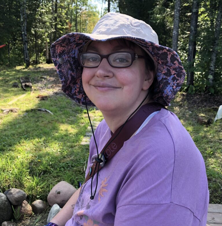 A woman wearing bucket hat and glasses smiles into camera wearing purple t-shirt in front of grassy outdoor background.