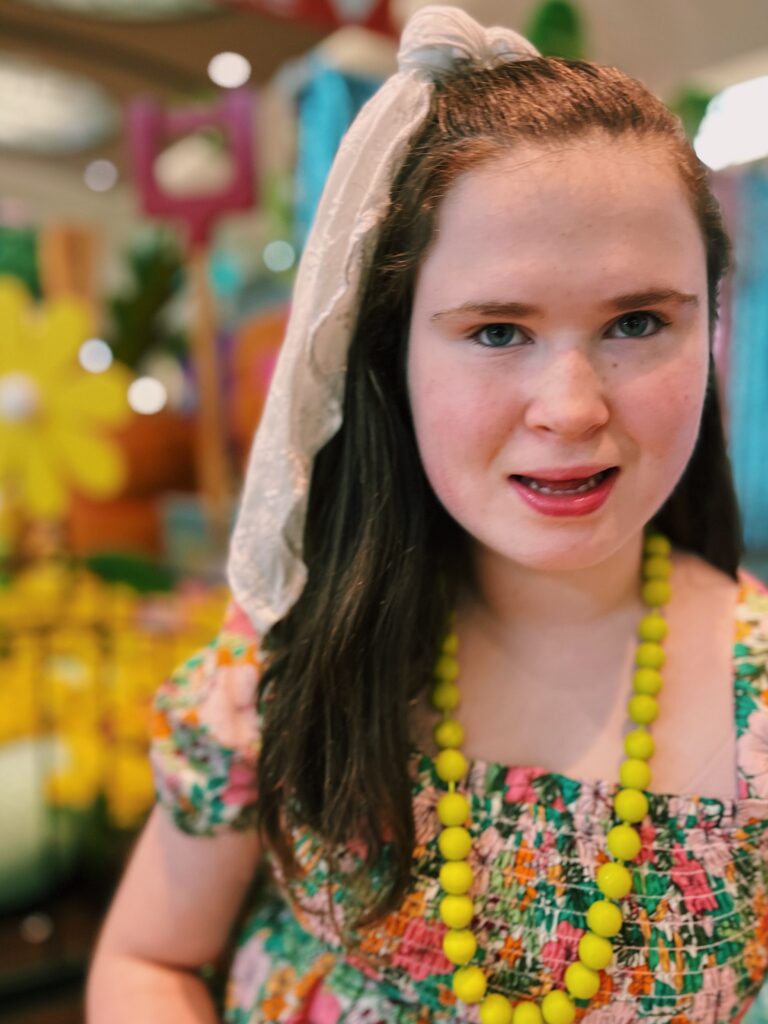 A young woman wearing brightly colored patterned dress and a yellow necklace smiles into camera with bright colors surrounding her.