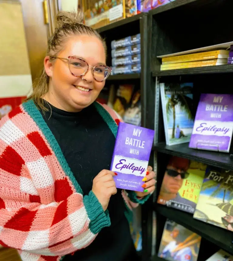 Woman wearing white and red checkered sweater smiles holding a purple book about epilepsy in a book store.