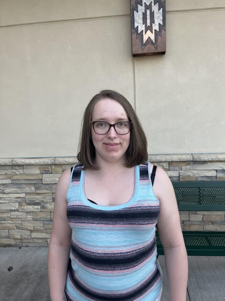 A woman wearing glasses and blue and black striped shirt smiles outside in front of brick building.