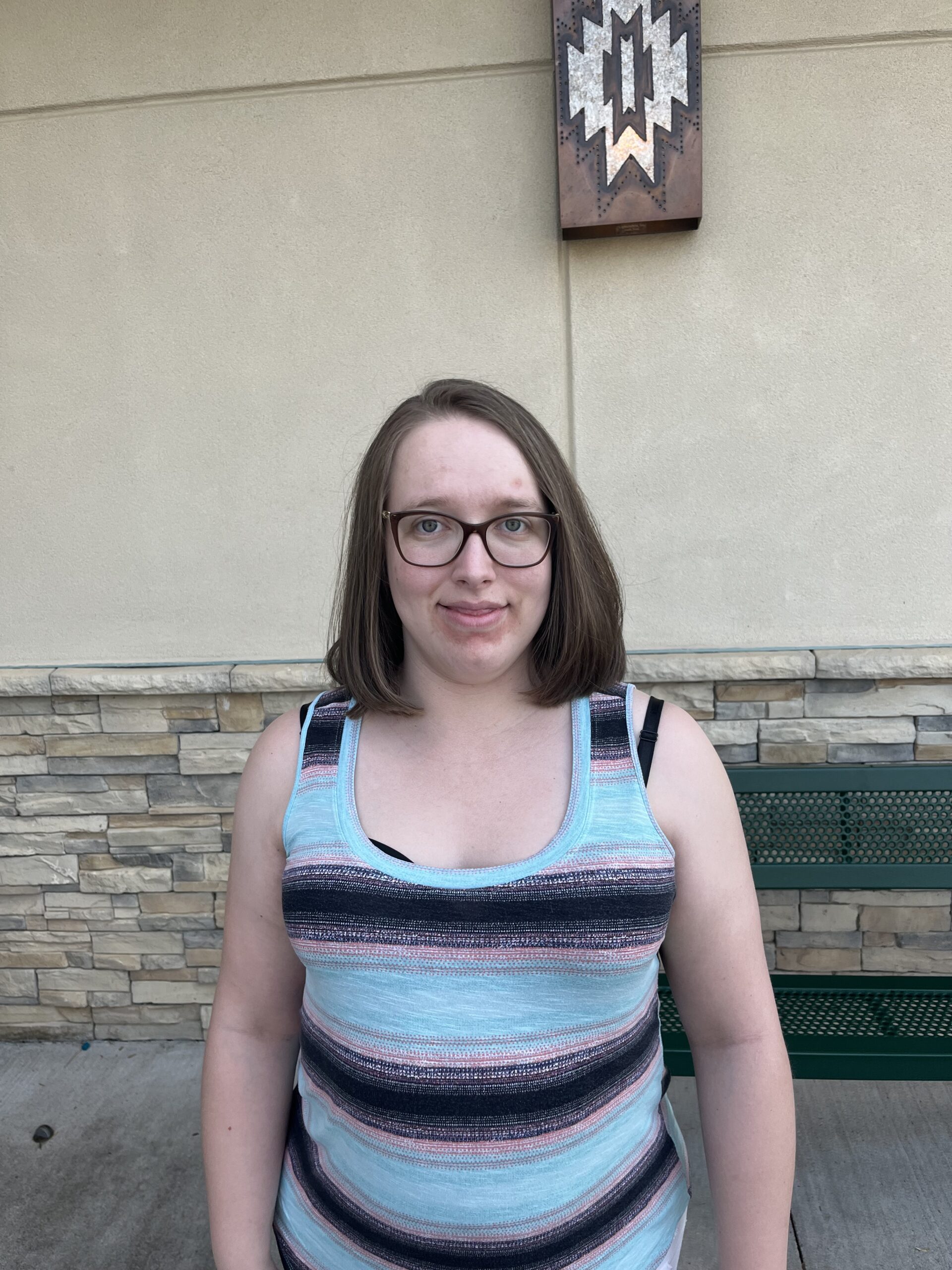 A woman wearing glasses and blue and black striped shirt smiles outside in front of brick building.