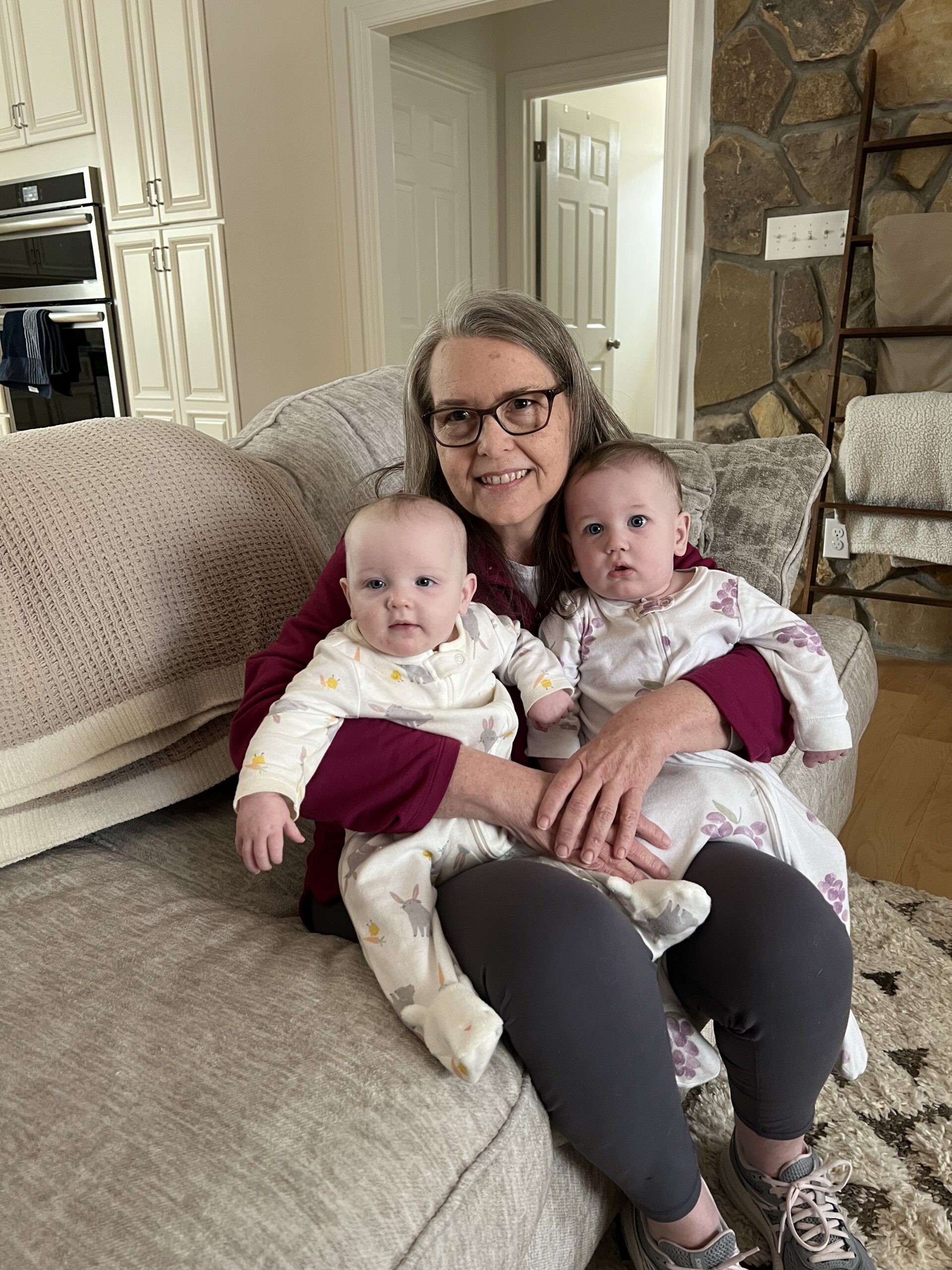 A woman holding two babies smiles at the camera while sitting on a couch.